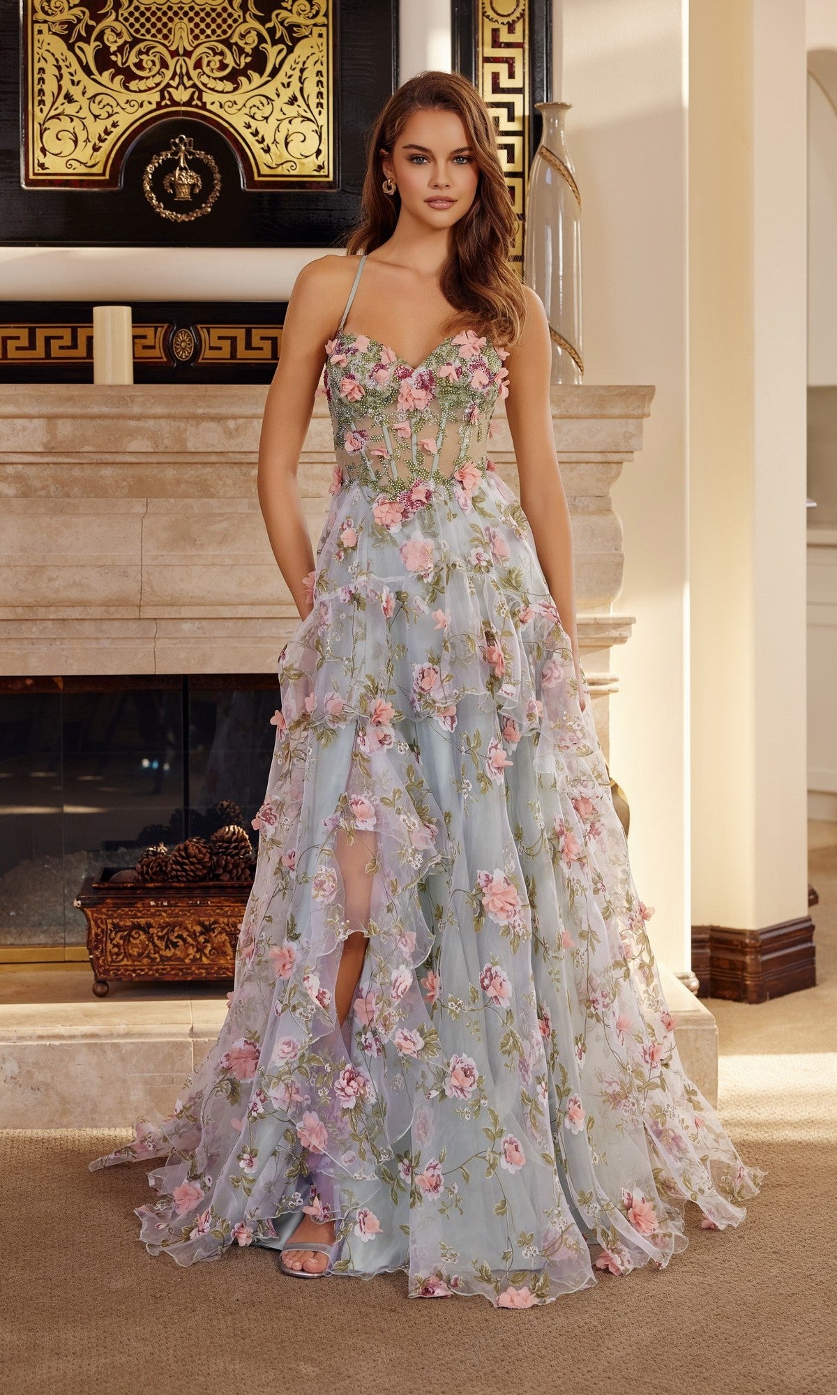 Indoors, a woman wears the Nox Anabel G1783 long prom dress—a sleeveless, floor-length gown with a structured bodice, sheer overlay, and 3D pink and green floral appliques.